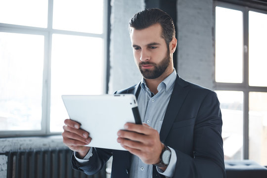 Working day...Serious bearded businessman in stylish suit and with branded watch on his hand is looking at digital tablet