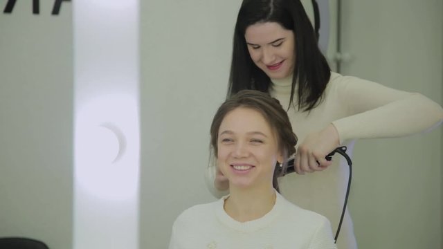 Beautiful woman with brown hair is having it treated with a curling iron by a hairdresser. Handheld real time establishing shot.