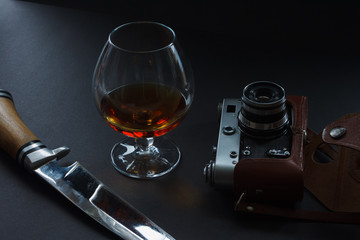 A glass of cognac,old analog film camera, hunting knife and pomegranate. Still life. Dark background