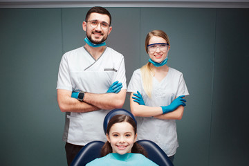 Nice positive male and female dentist with girl in dental chair. They look straight and smile. Adults hold hands crossed. Isolated on green background.