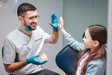 Male dentist giving high five to girl in dental chair. They are in room. Guy hold artificial jaw...