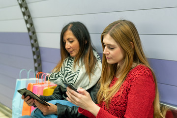 Couple of female friends with gift bags using smartphones seated in a subway station bench