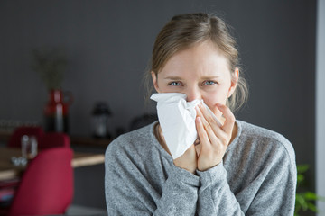 Unhappy sick woman blowing nose with napkin, looking at camera