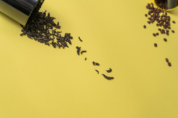 Large black tea leaves are scattered from black aluminum can in the foreground over a yellow surface, and roasted coffee beans are scattered from a yellow aluminum can in the background.