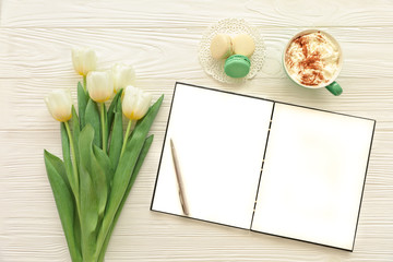 Beautiful bouquet of white tulips, empty open notebook, silver pen, cup of cappuccino, makaroons on the white wooden background.