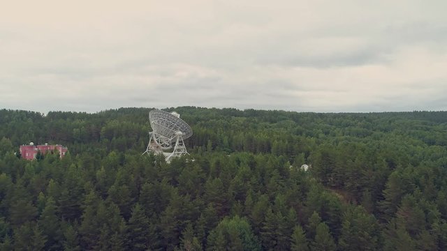 Aerial view shot of radio telescope in observatory. Backward flying. Move back. Green summer forest. Cloudy.