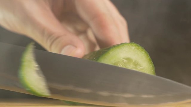 A man cuts with a knife a farmer's juicy and fresh cucumber in haze and evaporation, black background, close-up, slow motion