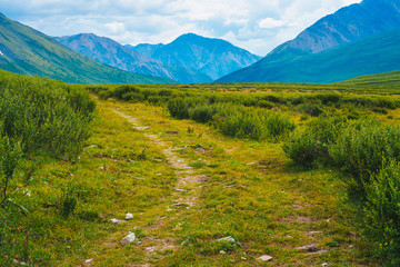 Spectacular view of distant giant mountains. Footpath through valley in highland in sunny day. Hiking path. Wonderful huge mountain range under cloudy sky. Amazing green landscape of majestic nature.