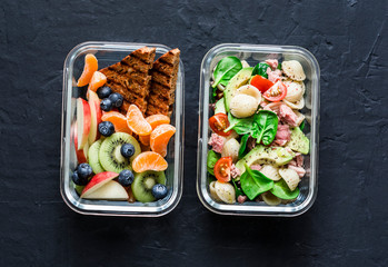 Office sweet and savory food lunch box. Pasta, tuna, spinach, avocado salad and fruit, peanut butter sandwiches lunch box on dark background top view