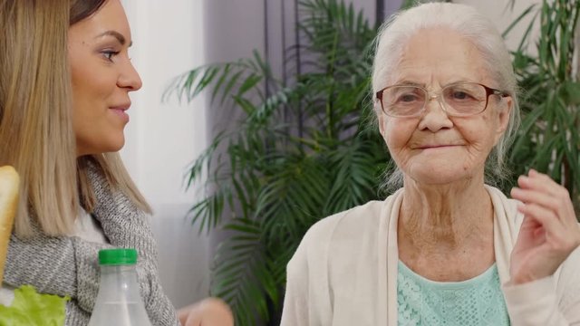 Tilt up shot of happy elderly grandmother putting on eyeglasses, embracing with young beautiful daughter holding bag of groceries and smiling at camera 