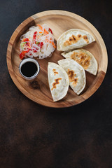 Wooden serving tray with fried potstickers, funchoza noodles salad and soy sauce. Flatlay on a dark brown metal surface with space
