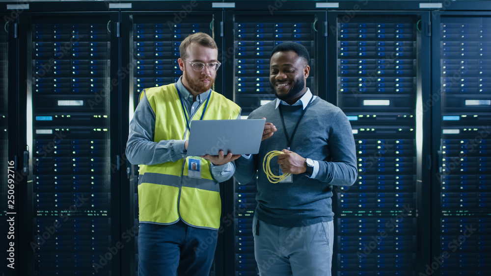 Poster Bearded IT Specialist in Glasses and High Visibility Vest with a Laptop Computer and Black Technician Colleague Talking in Data Center while Standing Next to Server Racks. Running Maintenance. 