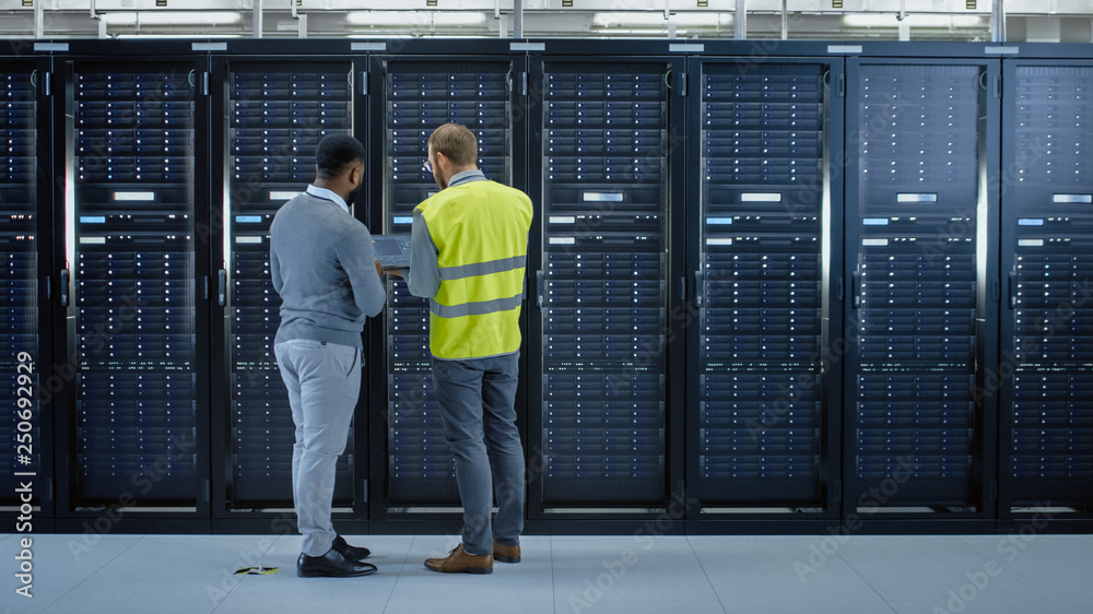 Poster Bearded IT Specialist in Glasses and High Visibility Vest with a Laptop Computer and Black Technician Colleague Talking in Data Center while Standing Next to Server Racks. Running Maintenance. 