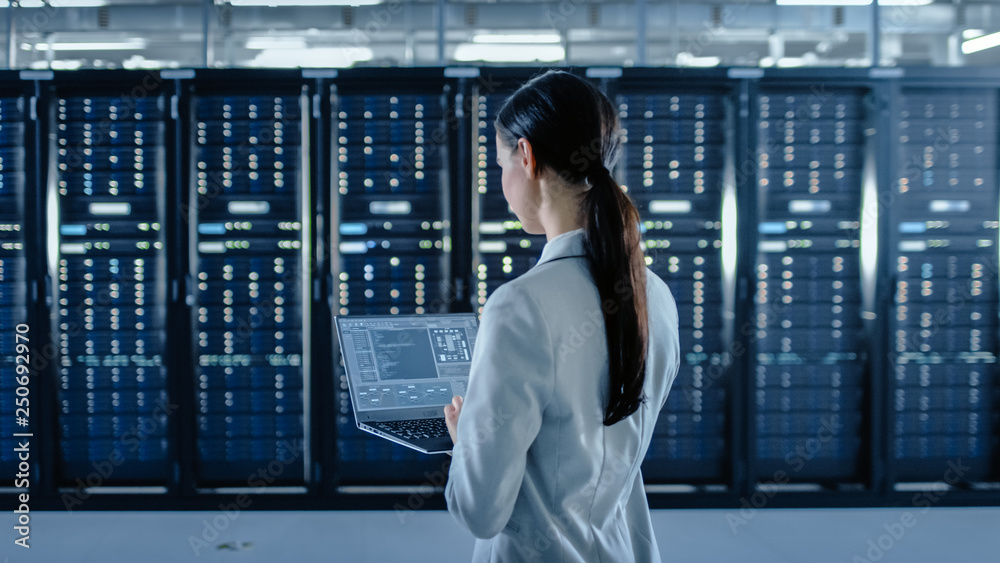 Poster back shot of a female data center it specialist works on laptop computer next to server rack corrido