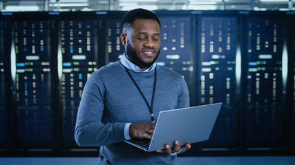 Black Data Center IT Technician Standing in the Middle of a Server Rack Corridor with a Laptop Computer. He is Visually Inspecting Working Server Cabinets.