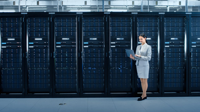 Female Data Center IT Engineer Works on Laptop Computer Next to Server Rack Corridor. Running Diagnostics or Doing Maintenance Work.