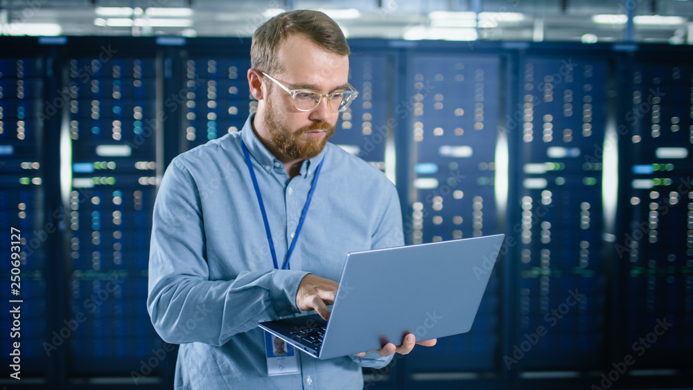 Fototapete bearded it specialist in glasses is working on laptop in data center next to server racks. running d