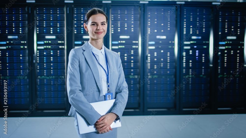 Poster female it specialist is standing in data center next to server racks, holding laptop and smiling to 