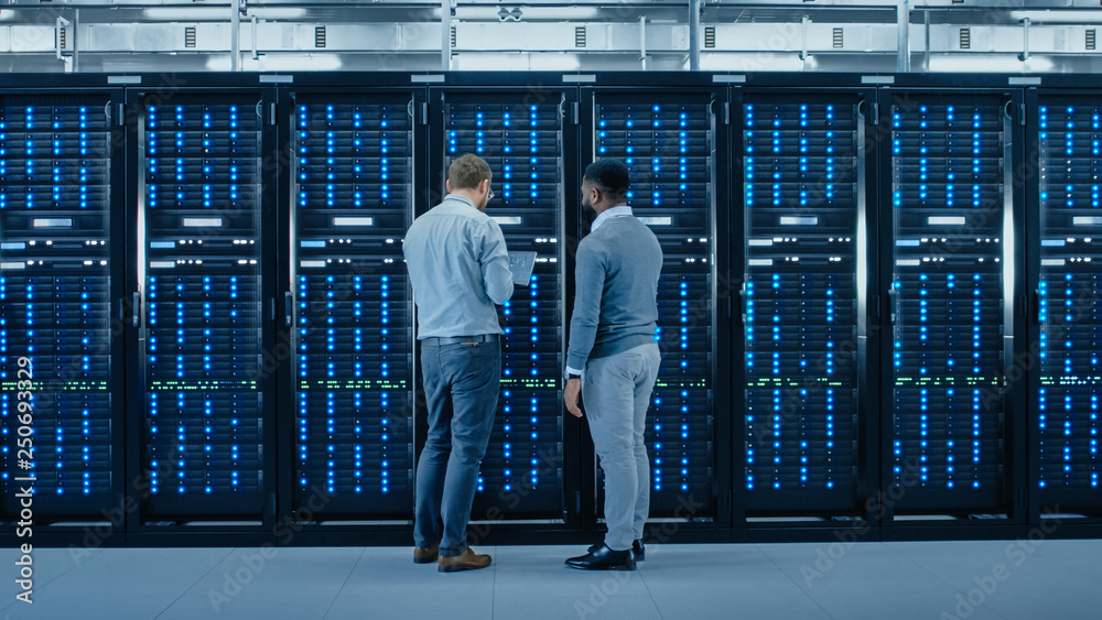 Poster Bearded IT Engineer in Glasses with a Laptop Computer and Black Technician Colleague Talking in Data Center while Standing Next to Server Racks. Running Diagnostics or Maintenance. 