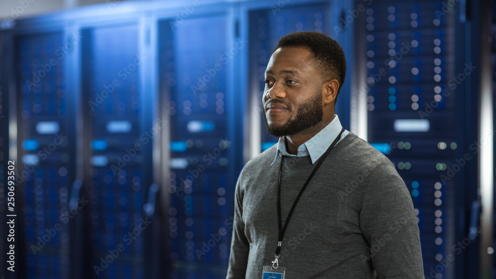 Poster Black Data Center IT Technician Standing in the Middle of a Server Rack Corridor. He is Smiling.