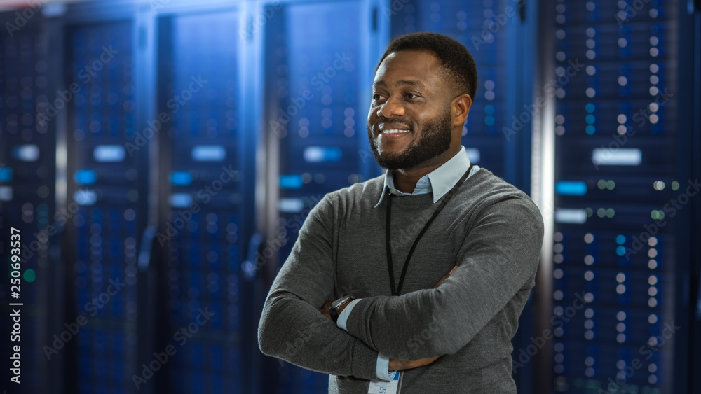 Poster Black Data Center IT Technician Standing in the Middle of a Server Rack Corridor. He is Standing with Crossed Arms and is Smiling.