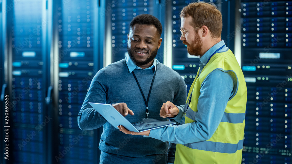 Poster Bearded IT Specialist in Glasses and High Visibility Vest with a Laptop Computer and Black Technician Colleague Talking in Data Center while Standing Next to Server Racks. Running Diagnostics. 