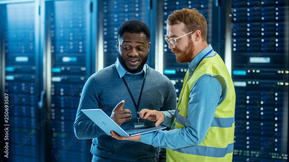Poster Bearded IT Specialist in Glasses and High Visibility Vest with a Laptop Computer and Black Technician Colleague Talking in Data Center while Standing Next to Server Racks. Running Diagnostics. 