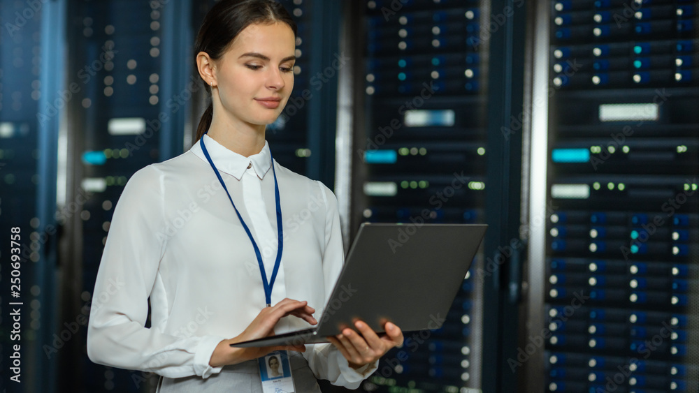 Poster Beautiful Data Center Female IT Technician Standing Near Server Rack Corridor with a Laptop Computer. She is Using Notebook and Working with Servers.