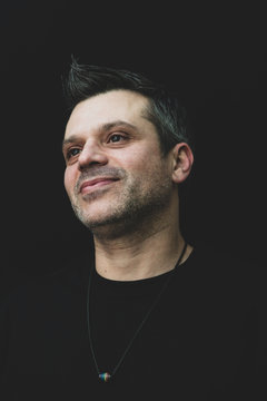 A gay man in black t shirt on a black background - wearing pride necklace, with salt and pepper hair and stubble. 