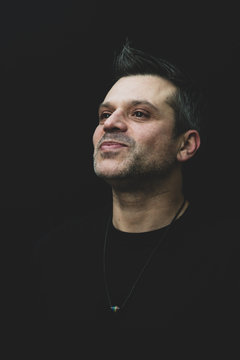 A Gay Man In Black T Shirt On A Black Background - Wearing Pride Necklace, With Salt And Pepper Hair And Stubble. 