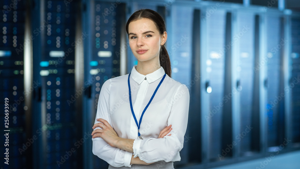 Poster female it specialist is standing at the camera in data center next to server racks and looking at th