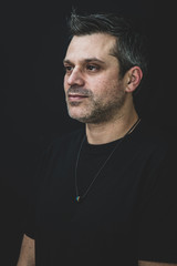 A gay man in black t shirt on a black background - wearing pride necklace, with salt and pepper hair and stubble. 