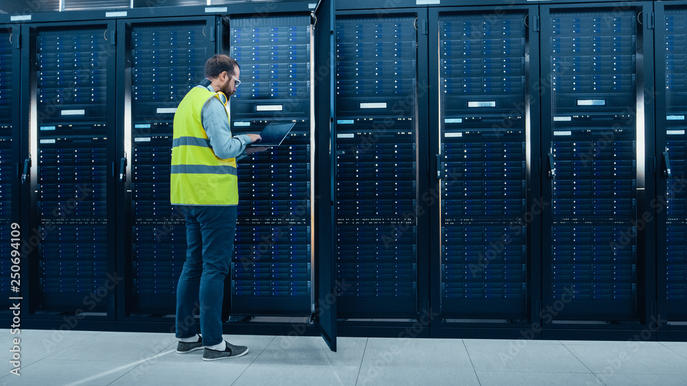 Leinwandbilder IT Engineer in High Visibility Vest is Working on Laptop in Data Center while Standing Before Server Rack. Running Diagnostics or Doing Maintenance Work.