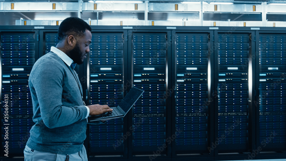 Aufkleber Black Data Center IT Technician Walking Through Server Rack Corridor with a Laptop Computer. He is Visually Inspecting Working Server Cabinets.