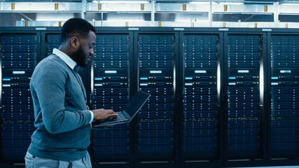 Black Data Center IT Technician Walking Through Server Rack Corridor with a Laptop Computer. He is...