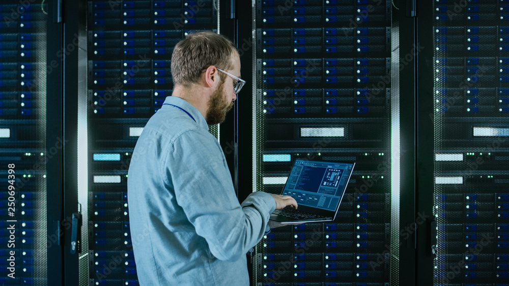 Fototapete Bearded Data Center IT Engineer in Glasses is Walking Through Server Rack Corridor with a Laptop Computer. He is Visually Inspecting Working Server Cabinets.