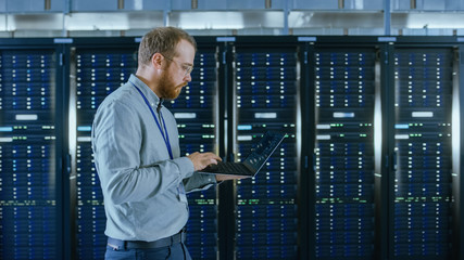 Bearded Data Center IT Engineer in Glasses is Walking Through Server Rack Corridor with a Laptop...