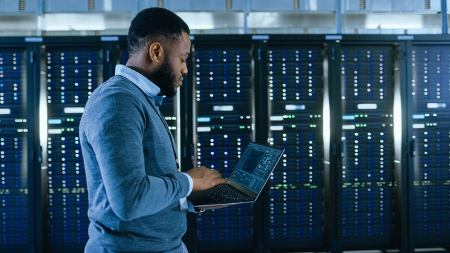 Black Data Center IT Technician Walking Through Server Rack Corridor with a Laptop Computer. He is Visually Inspecting Working Server Cabinets. - Powered by Adobe