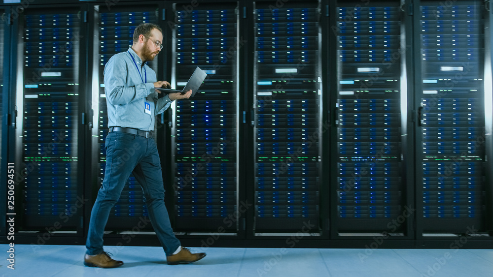 Aufkleber Bearded Data Center IT Professional Walking Through Server Rack Corridor with a Laptop Computer. He Stops and Wirelessly Inspects Working Server Cabinets.