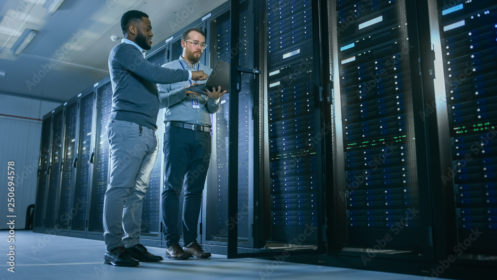 Poster Bearded IT Technician in Glasses with a Laptop Computer and Black Male Engineer Colleague are Talking in Data Center while Working Next to Server Racks. Running Diagnostics or Doing Maintenance Work. 