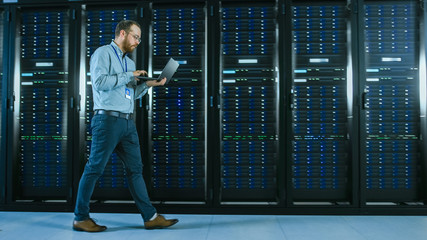 Bearded Data Center IT Professional Walking Through Server Rack Corridor with a Laptop Computer. He...