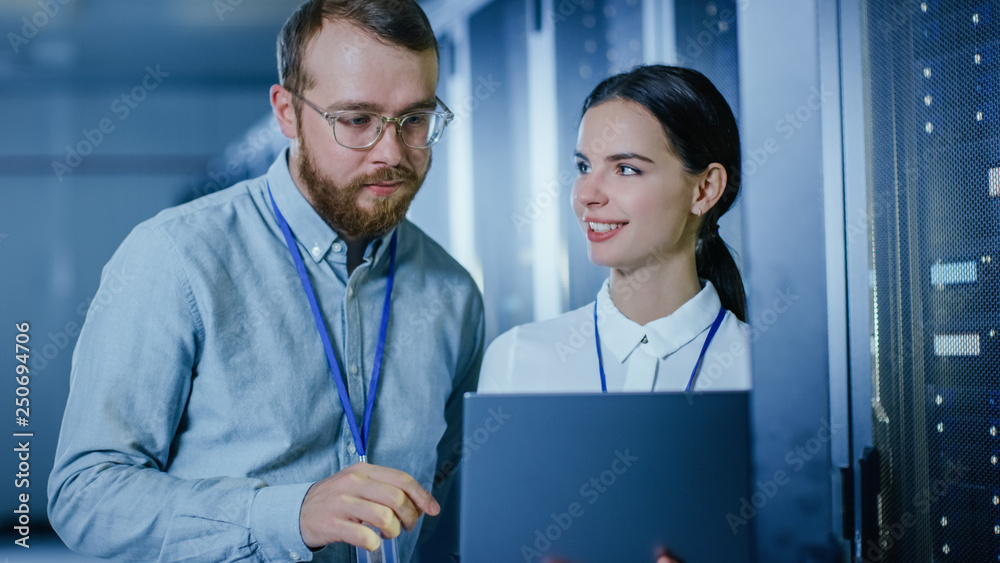 Poster Bearded IT Technician in Glasses and Beautiful Young Engineer Colleague with a Laptop Computer are Talking in Data Center while Working Next to Server Racks. Running Diagnostics or Doing Maintenance