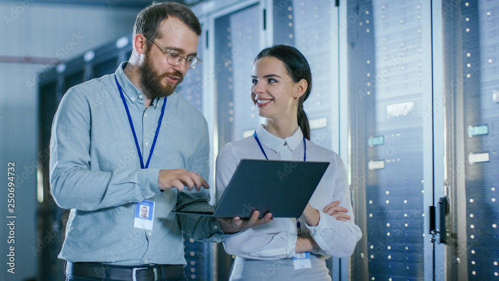 Aufkleber Bearded IT Technician in Glasses and Beautiful Young Engineer Colleague with a Laptop Computer are Talking in Data Center while Working Next to Server Racks. Running Diagnostics or Doing Maintenance