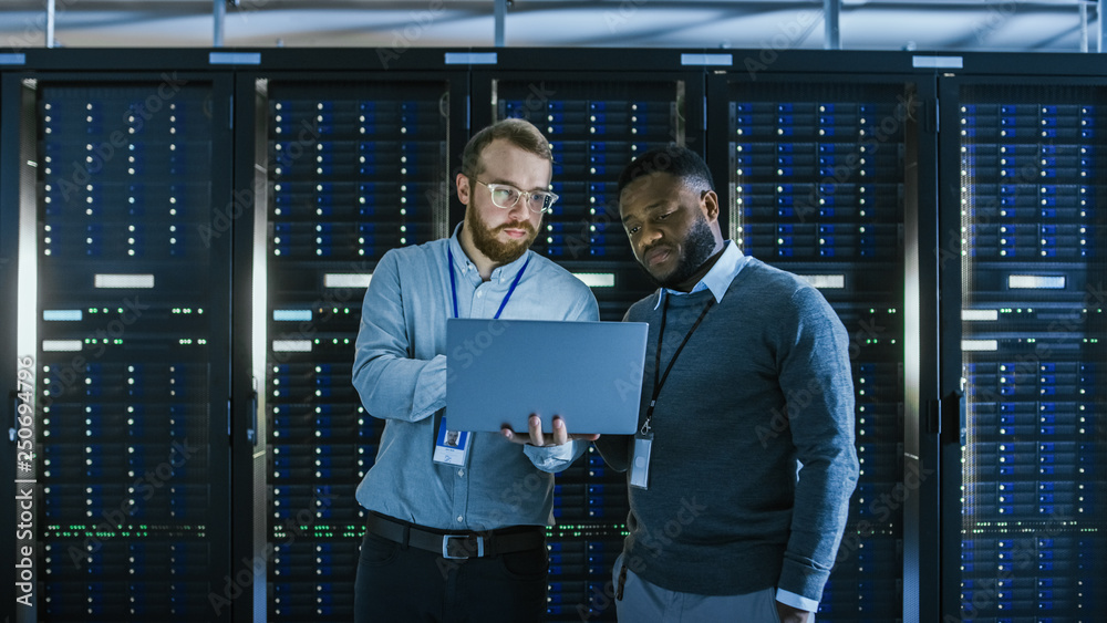 Aufkleber Bearded IT Technician in Glasses with a Laptop Computer and Black Male Engineer Colleague are Talking in Data Center while Working Next to Server Racks. Running Diagnostics or Doing Maintenance Work. 
