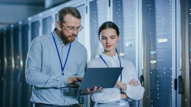 Bearded IT Technician in Glasses and Beautiful Young Engineer Colleague with a Laptop Computer are Talking in Data Center while Working Next to Server Racks. Running Diagnostics or Doing Maintenance