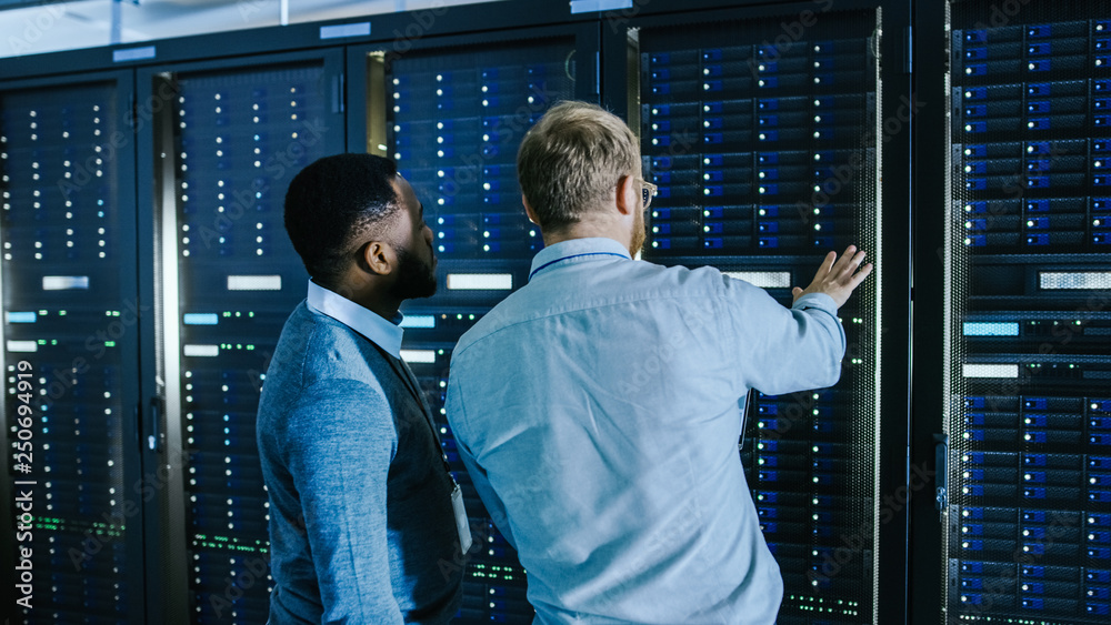 Poster Bearded IT Technician in Glasses with a Laptop Computer and Black Male Engineer Colleague are Talking in Data Center while Working Next to Server Racks. Running Diagnostics or Doing Maintenance Work. 