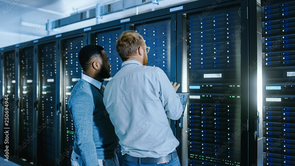 Poster Bearded IT Technician in Glasses with a Laptop Computer and Black Male Engineer Colleague are Talking in Data Center while Working Next to Server Racks. Running Diagnostics or Doing Maintenance Work. 