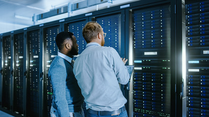 Bearded IT Technician in Glasses with a Laptop Computer and Black Male Engineer Colleague are Talking in Data Center while Working Next to Server Racks. Running Diagnostics or Doing Maintenance Work. 