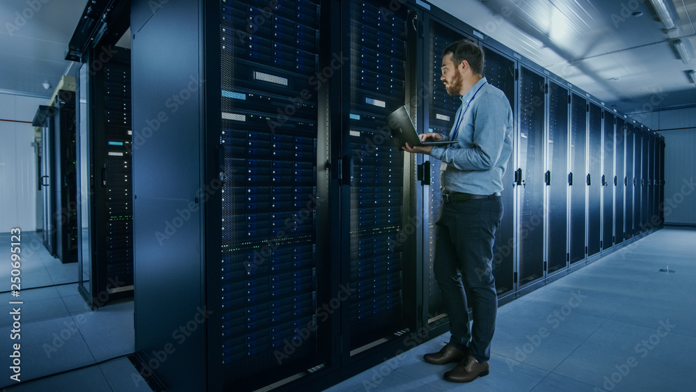 Poster Bearded IT Specialist in Glasses is Working on Laptop in Data Center while Standing Before Server Rack. Running Diagnostics or Doing Maintenance Work.