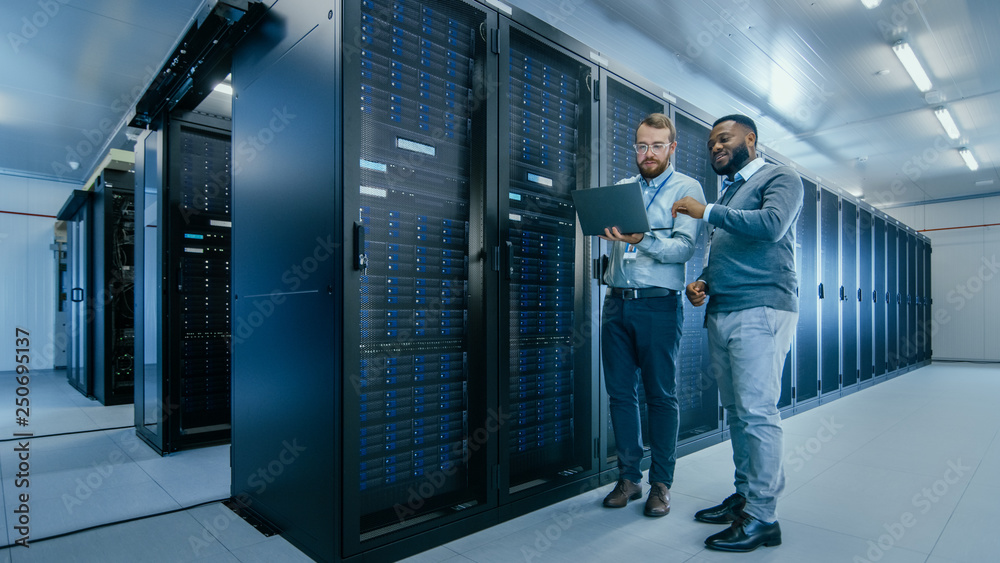 Aufkleber Bearded IT Technician in Glasses with a Laptop Computer and Black Male Engineer Colleague are Talking in Data Center while Working Next to Server Racks. Running Diagnostics or Doing Maintenance Work. 